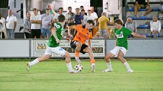 Drei Fußballspieler auf einem Feld, einer in Orange und zwei in Grün, mit einem Publikum, das hinter einem Zaun zusieht.