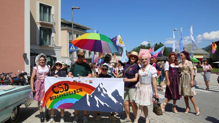 Eine Gruppe von Menschen in bunten Kostümen und mit einem Regenbogenbanner mit dem Text 'CSD Berchtesgaden' stehen vor einem Gebäude. Sie nehmen an einer Parade teil.