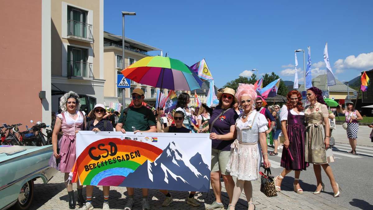 Eine Gruppe von Menschen in bunten Kostümen und mit einem Regenbogenbanner mit dem Text 'CSD Berchtesgaden' stehen vor einem Gebäude. Sie nehmen an einer Parade teil.