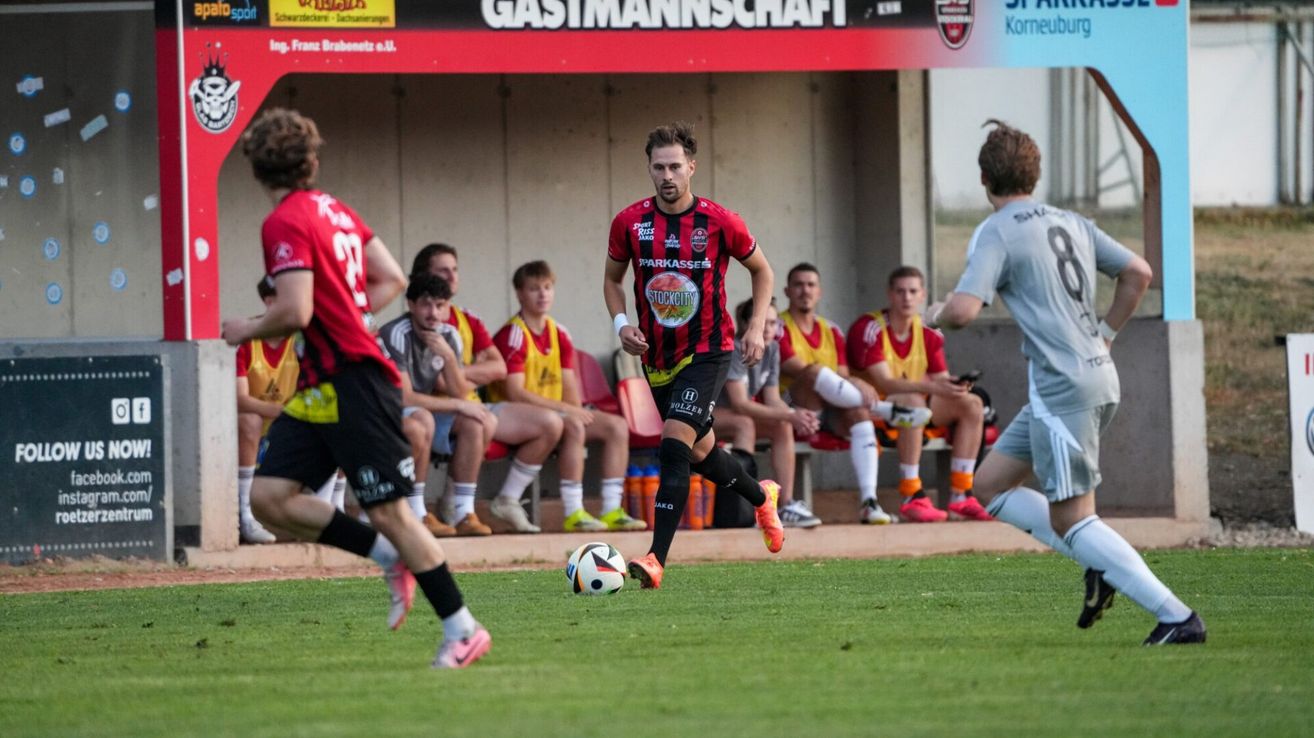Ein Fußballspieler in einem rot-schwarzen Trikot läuft und tritt einen Ball auf einem Feld, während andere Spieler auf einer Bank im Hintergrund sitzen.