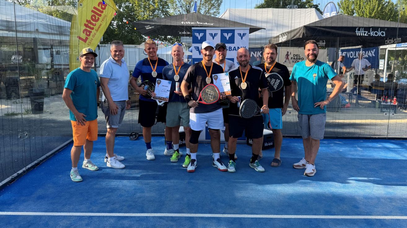 Eine Gruppe von Männern in Sportkleidung steht auf einem blauen Tennisplatz, hält Schläger und Medaillen. Sie lächeln und posieren für ein Foto.
