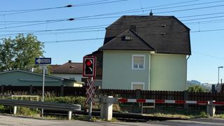 Eine rote Ampel mit einem X-Symbol steht neben einer Eisenbahnstrecke, mit einem Haus und einem Zaun im Hintergrund.