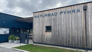 Das Äußere von Naturbad Pyh, ein Holzgebäude mit einem Schild, einem Fenster und einem Tor unter einem bewölkten Himmel.
