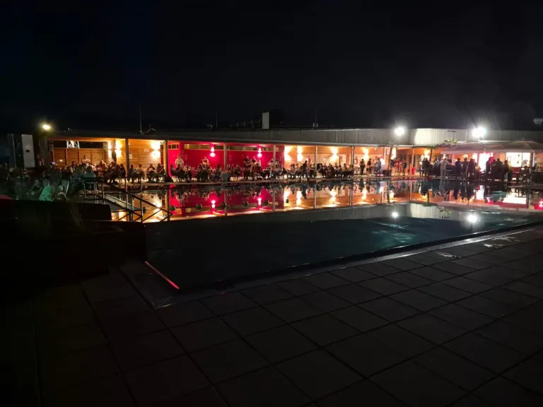 Ein nächtlicher Blick auf einen Poolbereich mit Menschen, die an Tischen sitzen, beleuchtet von roten Lichtern und umgeben von einem Gebäude mit roter Wand.