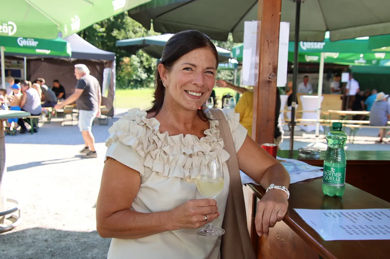 Eine Frau in einem weißen Rüschenkleid lächelt und hält ein Glas Weißwein bei einer Veranstaltung im Freien.