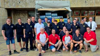 Eine Gruppe von Feuerwehrleuten in Uniform, einige kniend und andere stehend, posiert für ein Foto vor einem Gebäude, dessen LKW mit Wasserflaschen beladen ist und mit einer Plane abgedeckt ist.