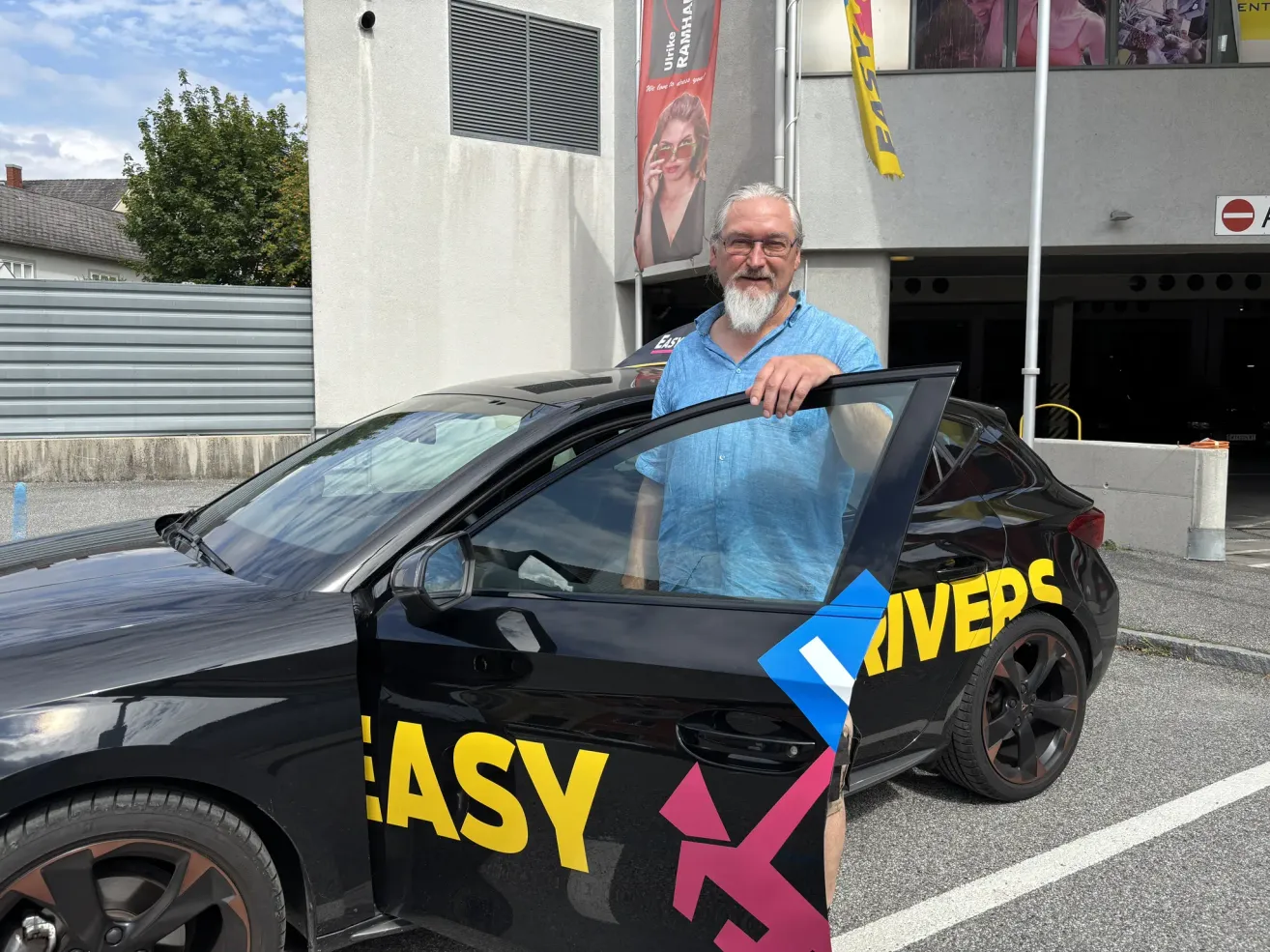 Ein älterer Mann mit weißem Bart steht neben einem schwarzen Auto mit dem Wort 'EASY' und einem Logo auf der Seite. Er trägt ein blaues Hemd und eine Brille. Hinter ihm steht ein Gebäude mit einem Banner, das eine Frau zeigt.