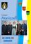 Eine Gruppe von fünf Personen in traditioneller Kleidung, die zusammenstehen, mit einem Hintergrund, der das Mariazell-Logo und das Wappen einer Gemeinde zeigt. Der Mann auf der linken Seite trägt eine Brille und einen Anzug, während die anderen traditionelle Kleidung tragen.