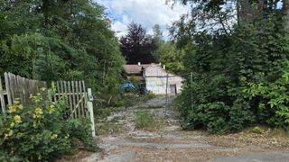 Ein Haus im Bau mit einem Holzzaun, einem blauen Bagger und einem Tor in einem bewaldeten Gebiet.