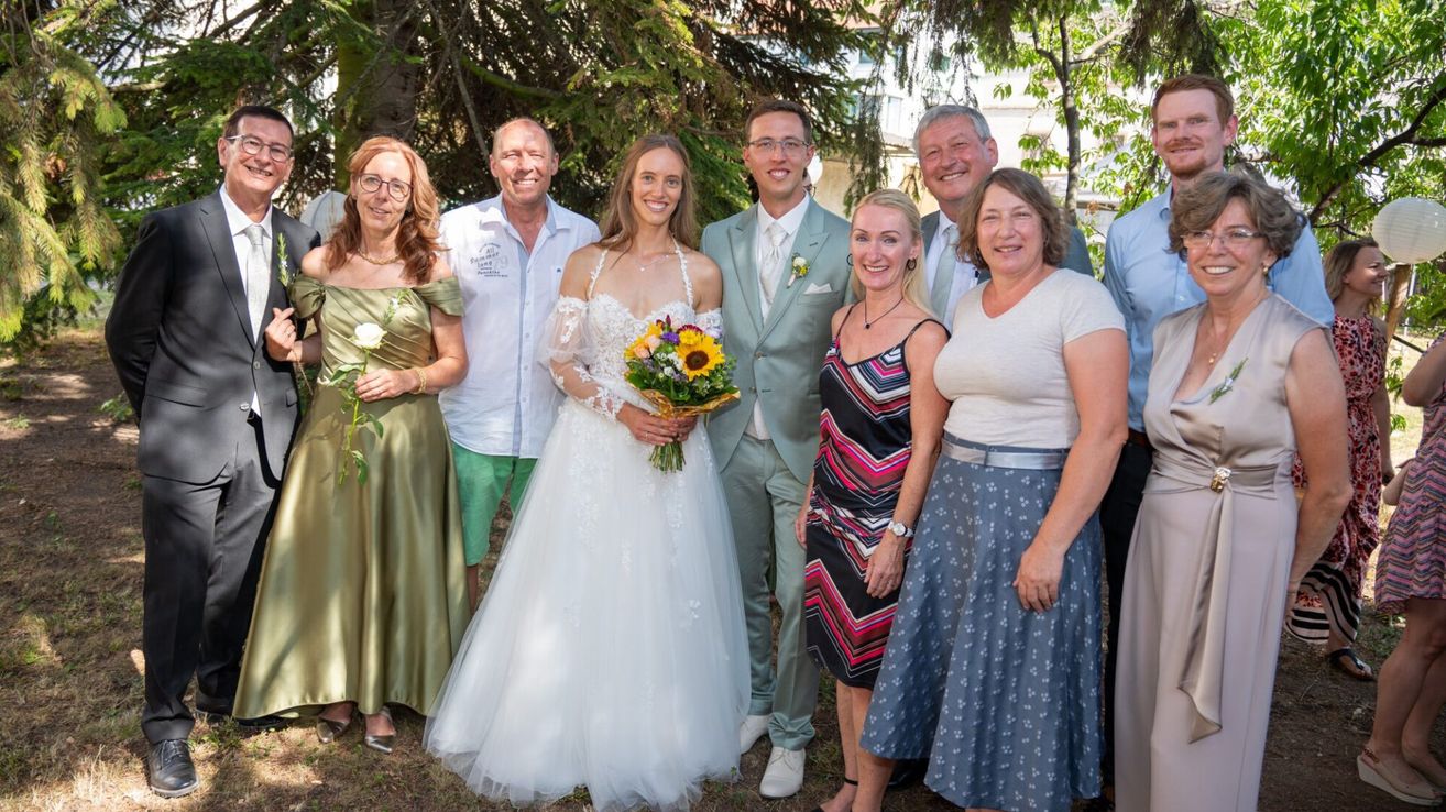 Eine Hochzeitsgesellschaft aus acht Personen, einschließlich der Braut und des Bräutigams, steht in einem Garten mit Bäumen im Hintergrund. Die Braut hält einen Strauß Sonnenblumen.