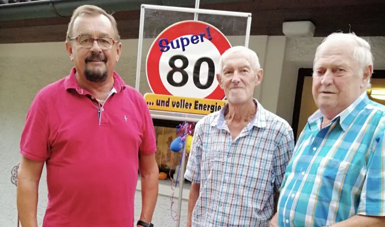 Two men stand in front of a sign that reads 'Super 80' with a red circle and yellow border. The man on the left wears a pink shirt, and the man on the right wears a checkered shirt.