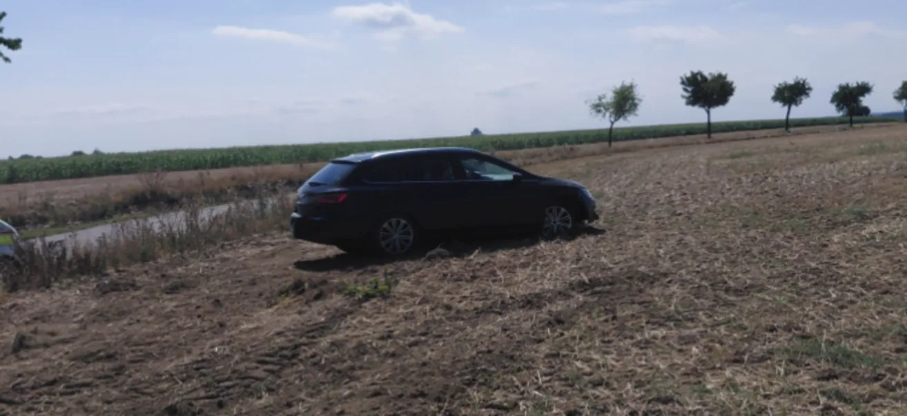 Ein schwarzes Auto steckt in einem schlammigen Feld fest, mit einem Baum im Hintergrund.