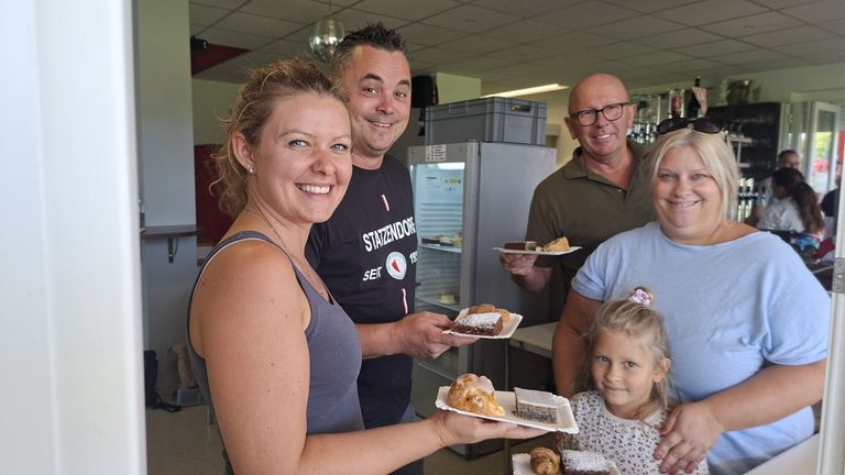 Eine Gruppe von Menschen, die lächeln und Teller mit Desserts in einer Küche halten. Eine Frau in einem grauen Tanktop hält zwei Teller. Hinter ihr hält ein Mann in einem schwarzen T-Shirt einen Teller. Ein anderer Mann in einem grünen Polohemd hält einen Teller. Ein Mädchen mit Zöpfen steht neben ihnen.