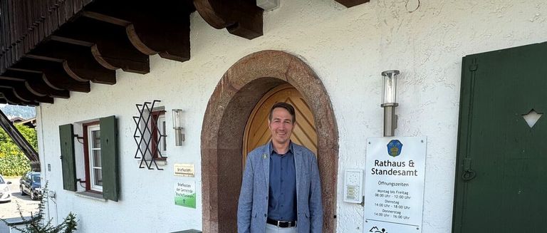 Ein Mann steht vor einem Gebäude mit einem Schild, auf dem Rathaus & Standesamt steht. Er trägt einen hellblauen Blazer und ein blaues Hemd. Das Gebäude hat eine Holztür und ein Fenster mit grünen Fensterläden. An der Wand befindet sich ein Schild mit dem Text Rathaus & Standesamt und den Öffnungszeiten.