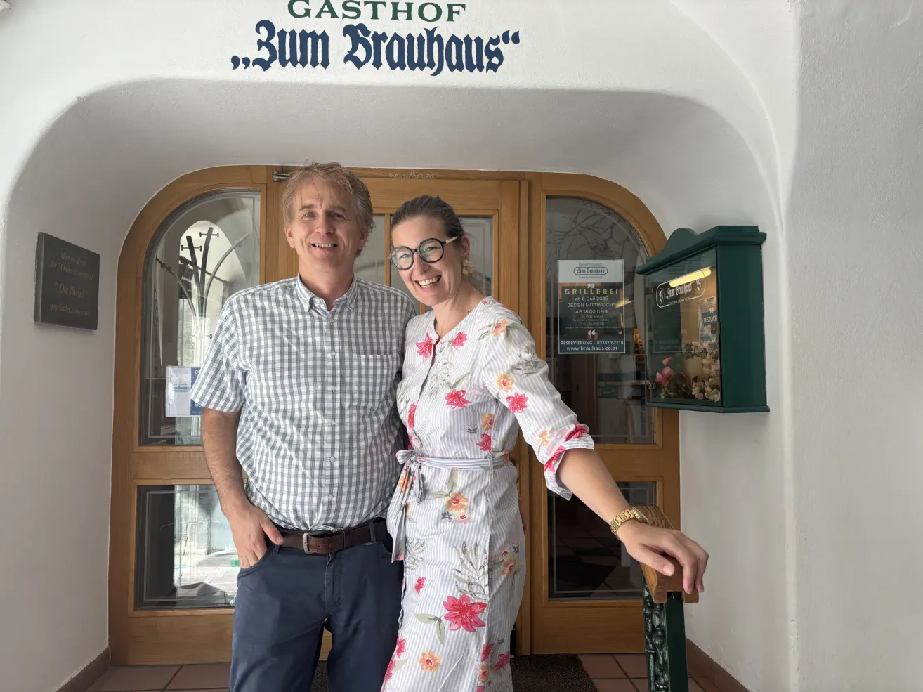 Ein Mann und eine Frau stehen vor einem Restaurant namens Gasthof zum Brauhaus, lächeln und posieren für ein Foto. Der Mann trägt ein kariertes Hemd und die Frau ein Blumenkleid. Hinter ihnen ist eine Holztür mit einem Schild zu sehen.