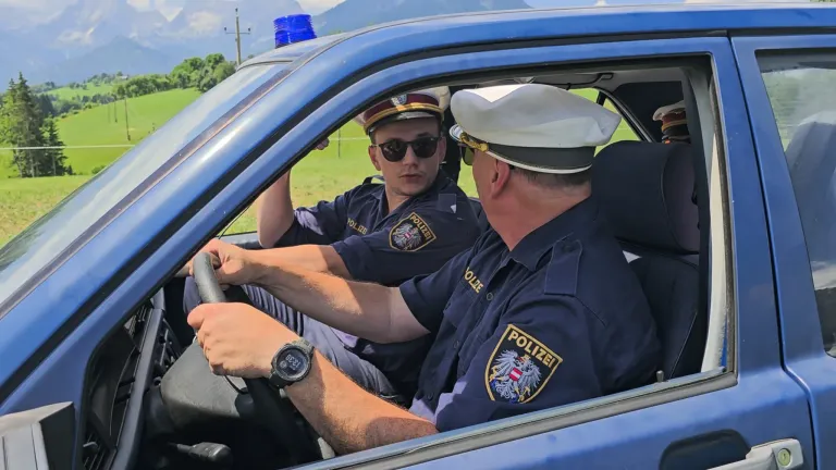 Two police officers in uniform sit in a blue car, one driving and the other with sunglasses on. The car has a police light on top.