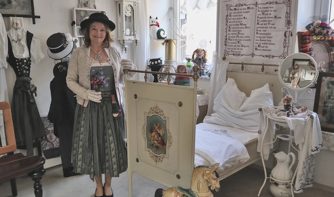 A woman in vintage attire stands next to a decorative crib with a horse toy in front of a window with various ornaments and a clock.
