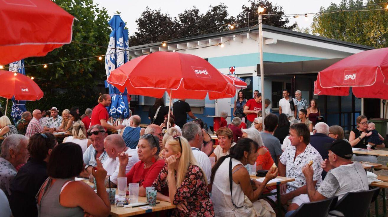 Eine Gruppe von Menschen sitzt unter Schirmen an Tischen in einem Outdoor-Essbereich. Ein roter Schirm mit dem Text 'SpO' ist sichtbar. Bäume und ein Gebäude mit einem roten Kreuzschild sind im Hintergrund.