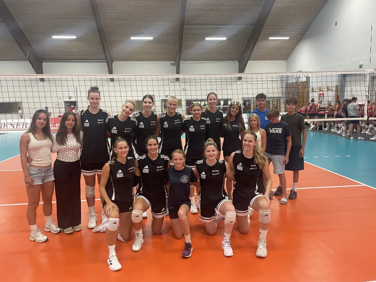 Eine Gruppe weiblicher Volleyballspielerinnen in schwarzen Uniformen posiert für ein Foto vor einem Netz auf einem Hallenplatz.
