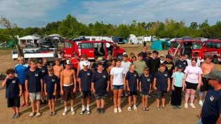 Eine Gruppe von Kindern und Erwachsenen steht vor einem roten Fahrzeug mit dem Wort 'VEHR' darauf, in einem Campingbereich mit Zelten und Bäumen im Hintergrund.