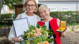 Zwei Frauen stehen lächelnd nebeneinander und halten einen Blumenstrauß und ein Zertifikat. Die Frau auf der linken Seite trägt eine Brille und ein weißes Oberteil, während die Frau auf der rechten Seite ein rotes ärmelloses Oberteil trägt und eine goldene Auszeichnung hält.