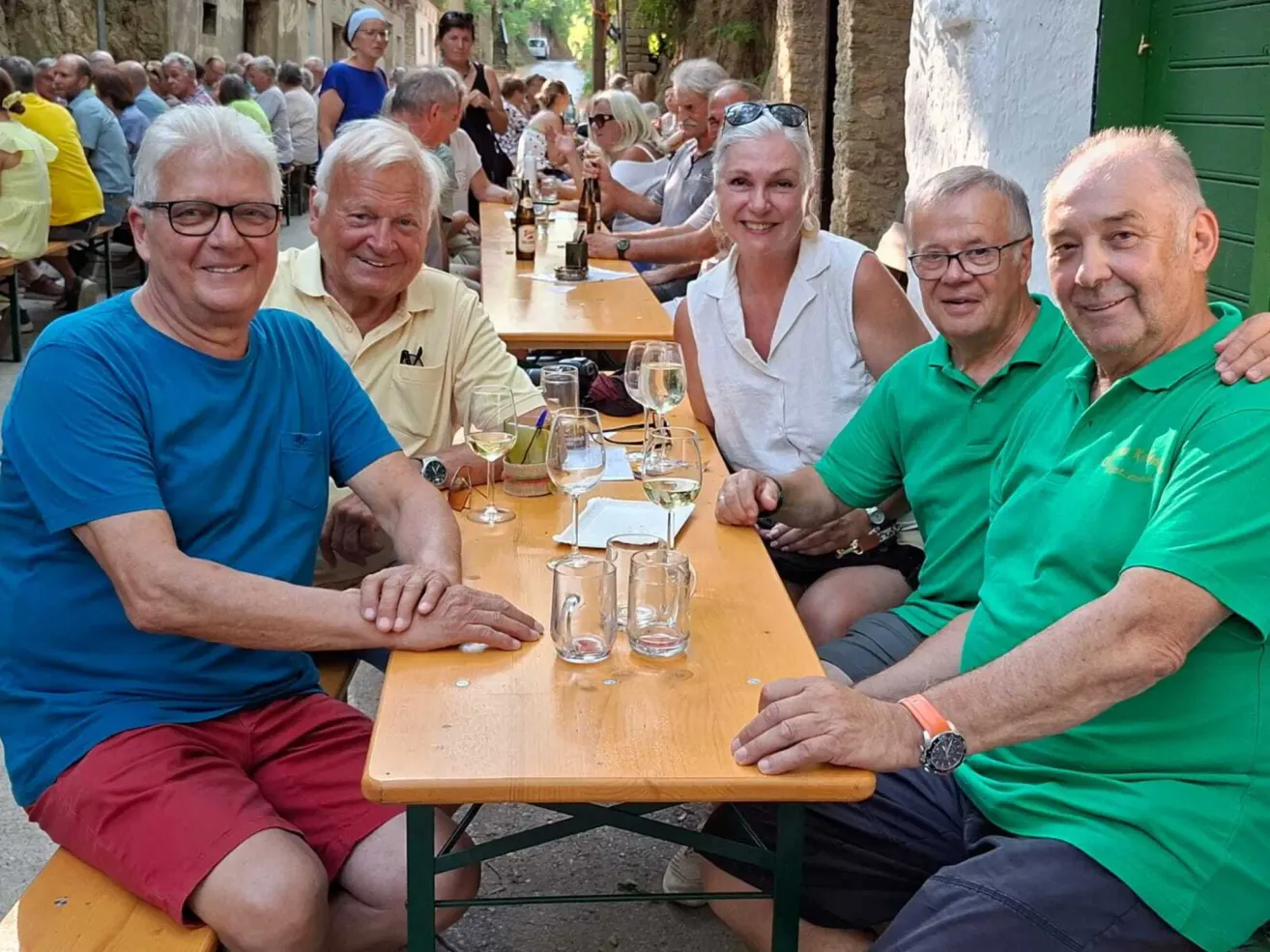 Eine Gruppe von Menschen, darunter vier Erwachsene, sitzt an einem Tisch in einer Außenumgebung, lächelt und posiert für ein Foto. Der Tisch ist mit Weingläsern, Tellern und Flaschen gedeckt. Im Hintergrund ist eine belebte Straße zu sehen, an der weitere Menschen an Tischen sitzen.