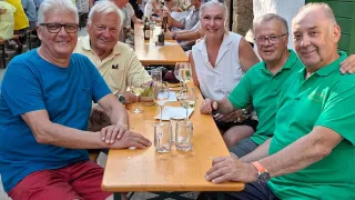 Eine Gruppe von Menschen, darunter vier Erwachsene, sitzt an einem Tisch in einer Außenumgebung, lächelt und posiert für ein Foto. Der Tisch ist mit Weingläsern, Tellern und Flaschen gedeckt. Im Hintergrund ist eine belebte Straße zu sehen, an der weitere Menschen an Tischen sitzen.