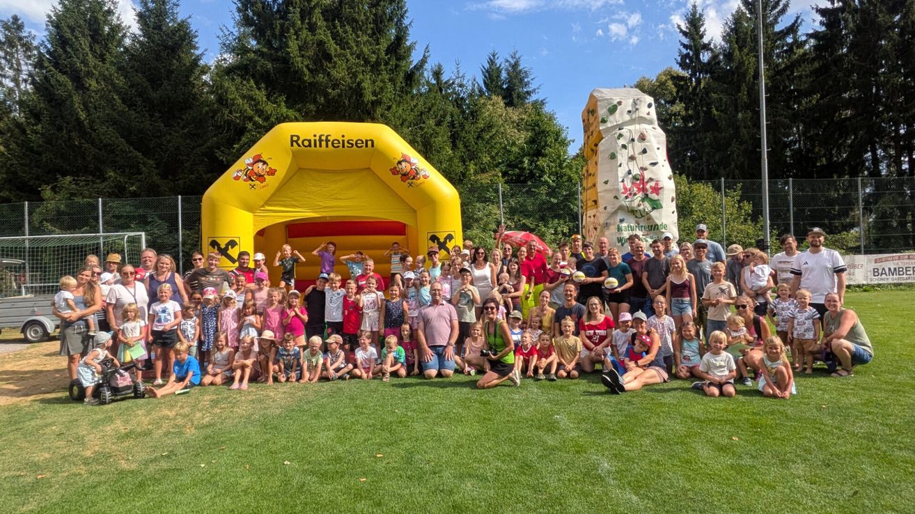Eine große Gruppe von Menschen, darunter Kinder und Erwachsene, posiert für ein Foto auf einem Rasenfeld mit einem Raiffeisen-Aufblasbaren und einer Kletterwand im Hintergrund.