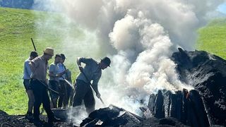 Eine Gruppe von Männern arbeitet an einem Feuerring auf einer Wiese, wobei ein Mann einen Schaufel benutzt, um Kohlen zu verteilen.