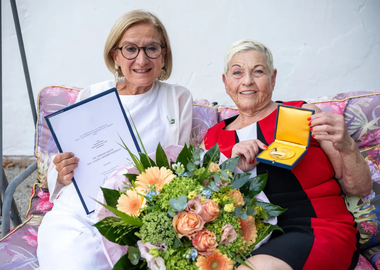 Zwei Frauen sitzen auf einer Couch, eine hält ein Zertifikat und die andere eine Medaille. Sie lächeln beide und posieren für ein Foto.