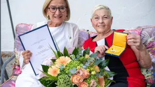 Zwei Frauen sitzen auf einer Couch, eine hält ein Zertifikat und die andere eine Medaille. Sie lächeln beide und posieren für ein Foto.