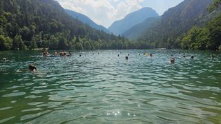 Eine Gruppe von Menschen schwimmt in einem See, umgeben von Bergen und Bäumen an einem sonnigen Tag.