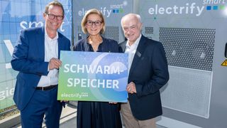 Drei Personen stehen zusammen und halten ein Schild mit der Aufschrift 'SCHWARZ-SPEICHER' vor einem Hintergrund mit dem Wort 'electrify'. Der Mann links trägt eine Brille und einen Gürtel, die Frau in der Mitte hat eine Brille und einen blauen Mantel, und der Mann rechts trägt einen Anzug.