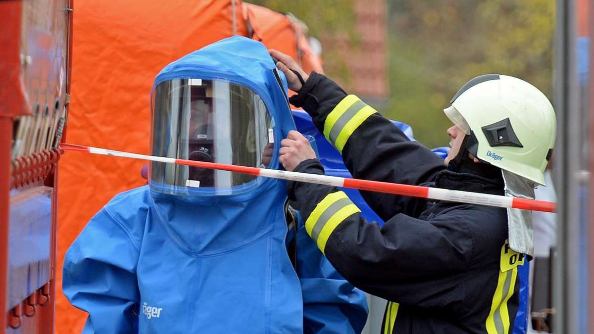 Ein Feuerwehrmann in voller Schutzausrüstung hilft einem Kollegen, einen blauen Gefahrgutanzug anzulegen.