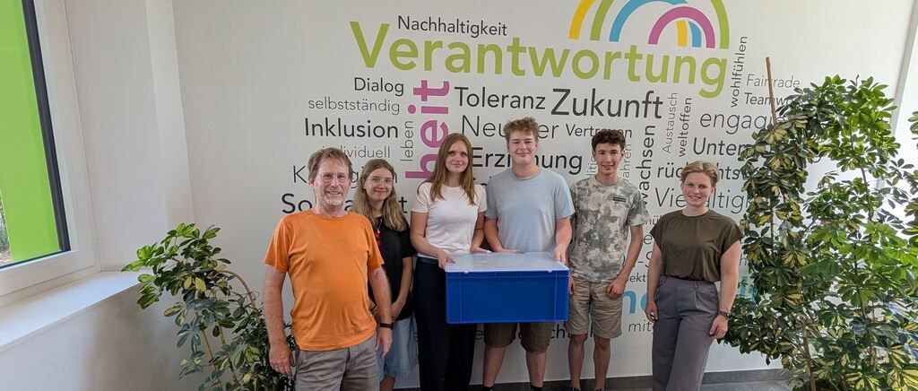 Eine Gruppe von Menschen steht vor einer Wand mit Wörtern wie 'Verantwortung' und 'Nachhaltigkeit'. Sie halten eine blaue Box.