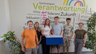 Eine Gruppe von Menschen steht vor einer Wand mit Wörtern wie 'Verantwortung' und 'Nachhaltigkeit'. Sie halten eine blaue Box.