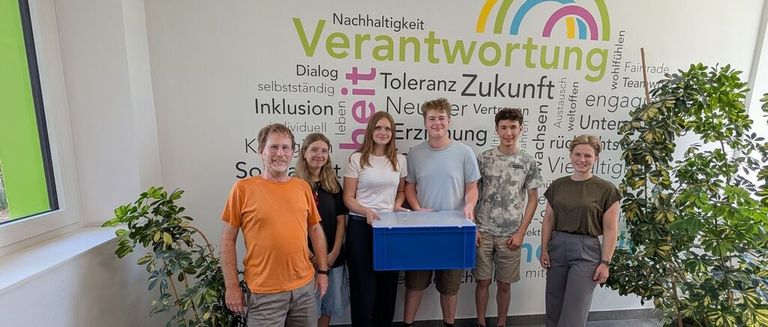 Eine Gruppe von Menschen steht vor einer Wand mit Wörtern wie 'Verantwortung' und 'Nachhaltigkeit'. Sie halten eine blaue Box.