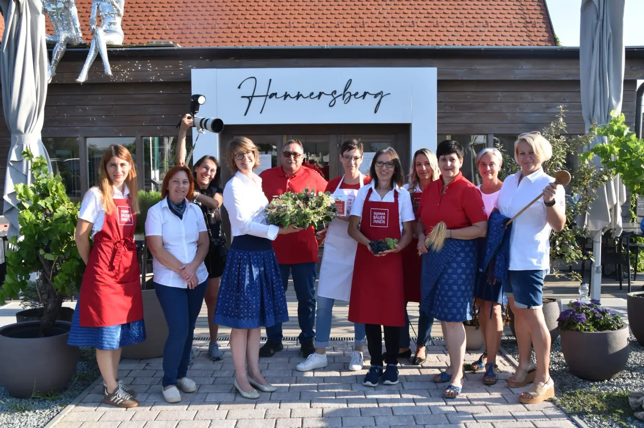 A group of women and men in red aprons stand outside a building named Hannerberg, holding flowers and vegetables, with a photographer capturing the moment.