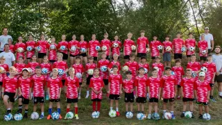 Eine Gruppe junger Fußballspieler in roten und weißen Trikots, die Fußballbälle halten und für ein Teamfoto vor einem von Bäumen gesäumten Hintergrund posieren.