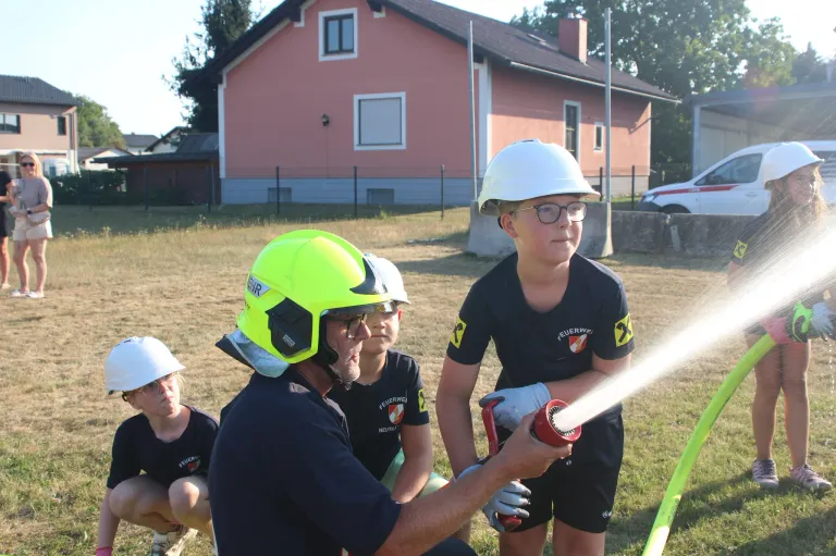Eine Gruppe von Kindern in Feuerwehruniformen übt das Benutzen eines Feuerlöschers unter Anleitung eines Erwachsenen auf einem Feld.