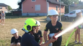 Eine Gruppe von Kindern in Feuerwehruniformen übt das Benutzen eines Feuerlöschers unter Anleitung eines Erwachsenen auf einem Feld.