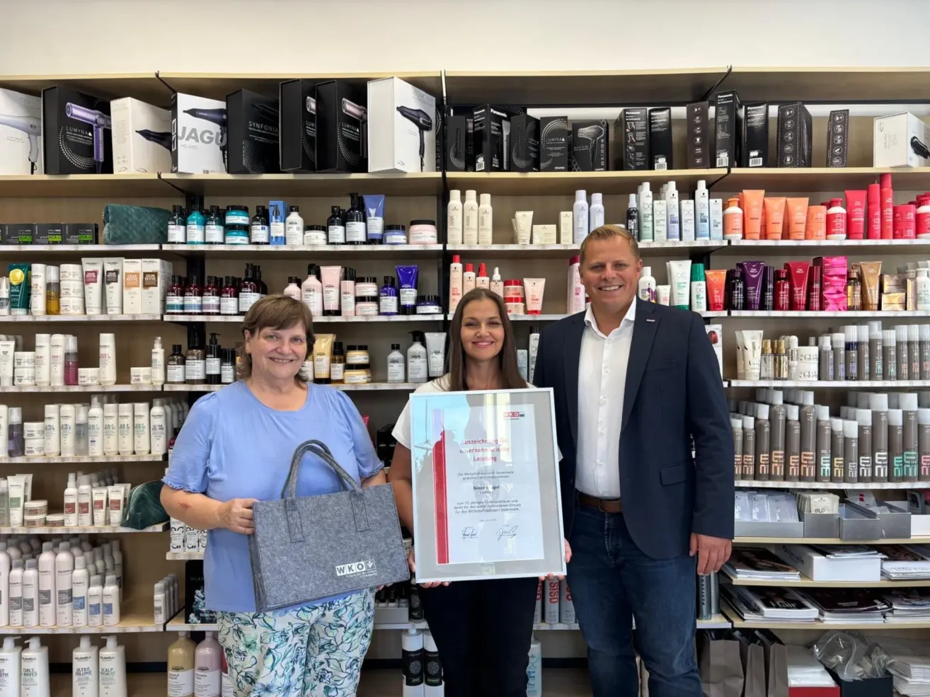 Drei Personen stehen vor einem Regal voller Haarprodukte, wobei eine Frau ein gerahmtes Zertifikat und eine Handtasche hält.