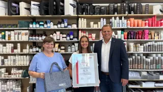 Drei Personen stehen vor einem Regal voller Haarprodukte, wobei eine Frau ein gerahmtes Zertifikat und eine Handtasche hält.
