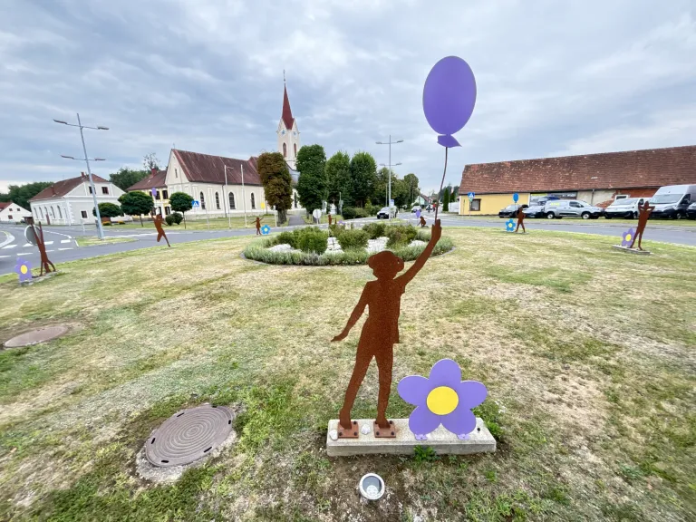 A silhouetted figure holding a purple balloon stands in a grassy field, with a flower beside it, in front of a church and a road.