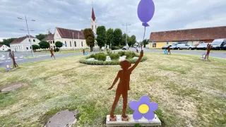 Eine silhouettierte Figur, die einen lila Luftballon hält, steht auf einem Rasen, mit einer Blume daneben, vor einer Kirche und einer Straße.