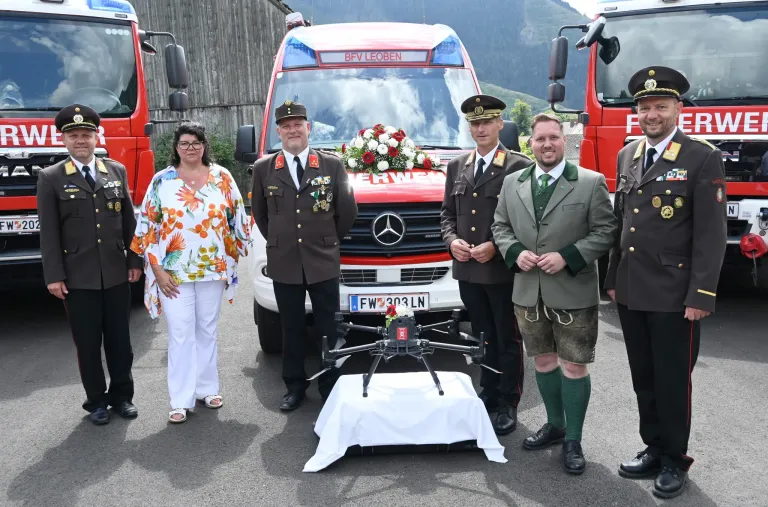 A group of people in uniforms stand next to a fire truck with flowers and a drone on a table in front of them.