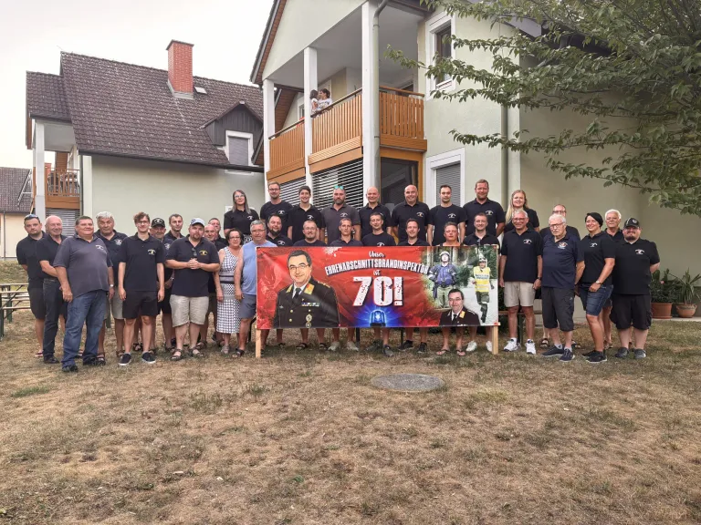 Eine Gruppe von Menschen in Uniform und Freizeitkleidung posiert für ein Foto vor einem Haus mit einem Banner, das 70 Jahre Dienst feiert.