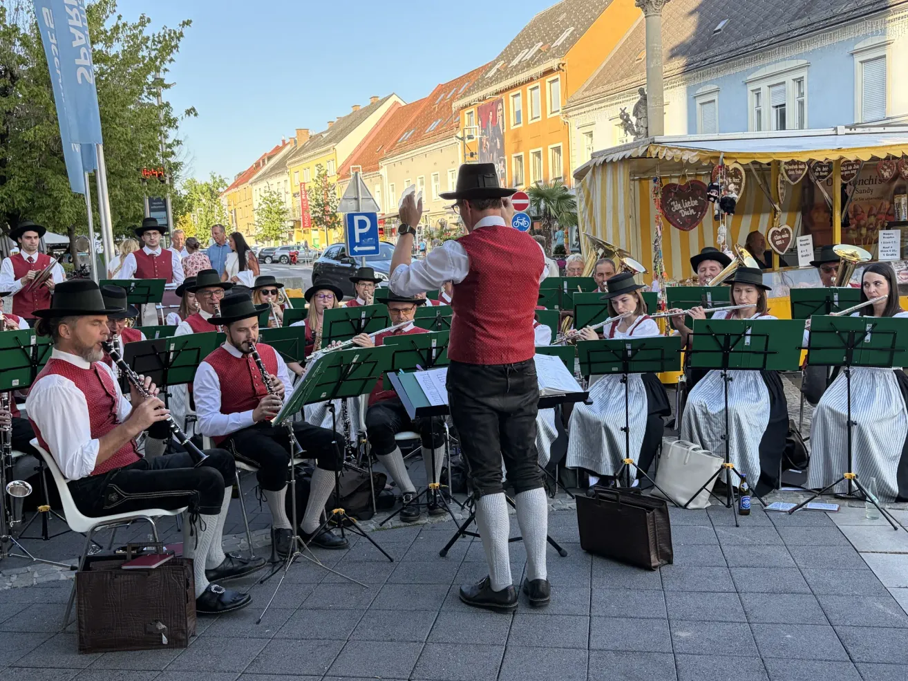 Eine Straßenband spielt im Freien, ein Dirigent leitet, Musiker spielen verschiedene Instrumente und ein Publikum schaut zu.