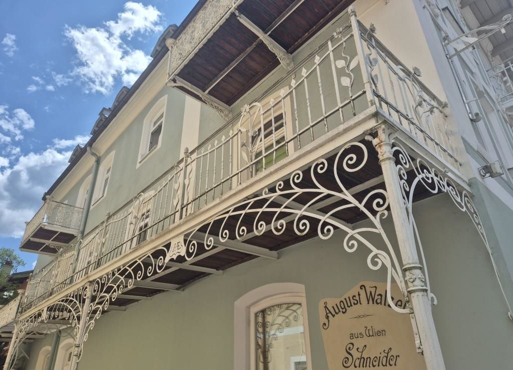 Ein altes Gebäude mit einem Balkon, der mit kunstvollen Metallgittern und einem Schild, das August Walcher aus Wien Schneider liest, verziert ist.
