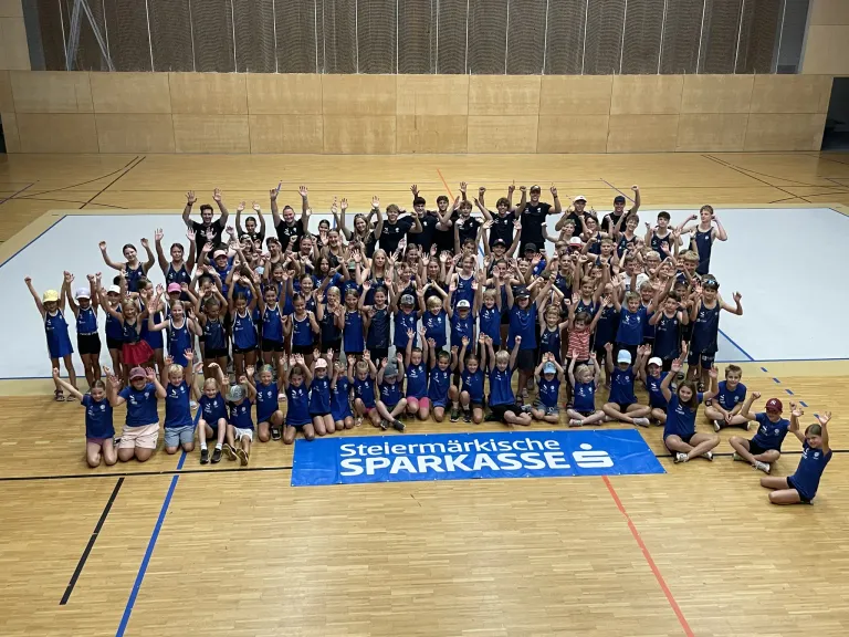 Eine Gruppe von Kindern und Erwachsenen in blauen Uniformen posiert für ein Foto in einer Turnhalle mit einem Banner, das 'Steiermarkische Sparkasse' steht.